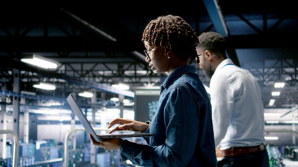 junge Frau im Vordergrund mit Laptop, dahinter junger Mann, IT Arbeitsumfeld