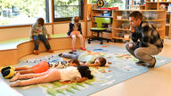 Szene einer Kinderbetreuung: Kinder kommen liegend oder sitzend zur Ruhe unter der Aufsicht eines Erwachsenen