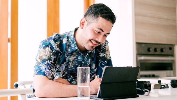 indonesischer Mann sitzt an einem Tisch, schaut auf ein Laptop und lächelt