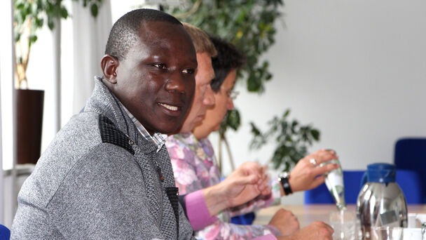 afrikanischer Mann sitzt mit anderen in einer Tischreihe bei einem Seminar und blickt in die Kamera