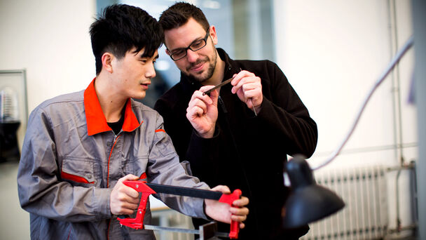 zwei Männer in Arbeitskleidung sehen sich ein Sägeblatt an, ein mutmaßlich chinesischer Mann arbeitet mit einer Handsäge
